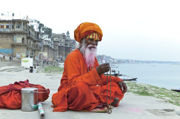 Homme sage en orange méditant près de l'eau, avec une barbe blanche et des perles.