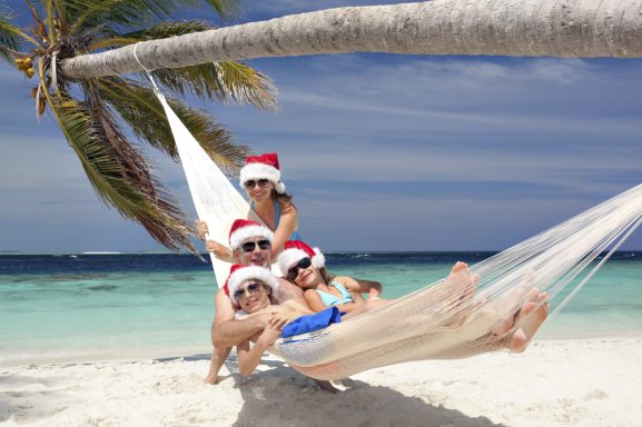 Quatre personnes sur un hamac sur une plage tropicale, portant des chapeaux de Noël.